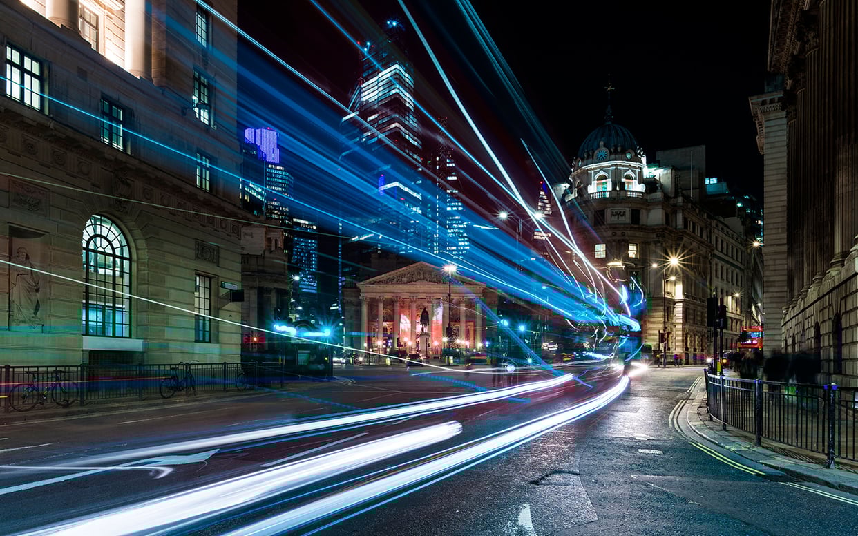 Dynamic long-exposure shot of city traffic at night showing vibrant blue and white light trails across a bridge.