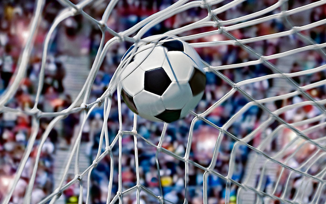 Close-up of a football hitting the back of a net during a match with blurred stadium lights in the background.