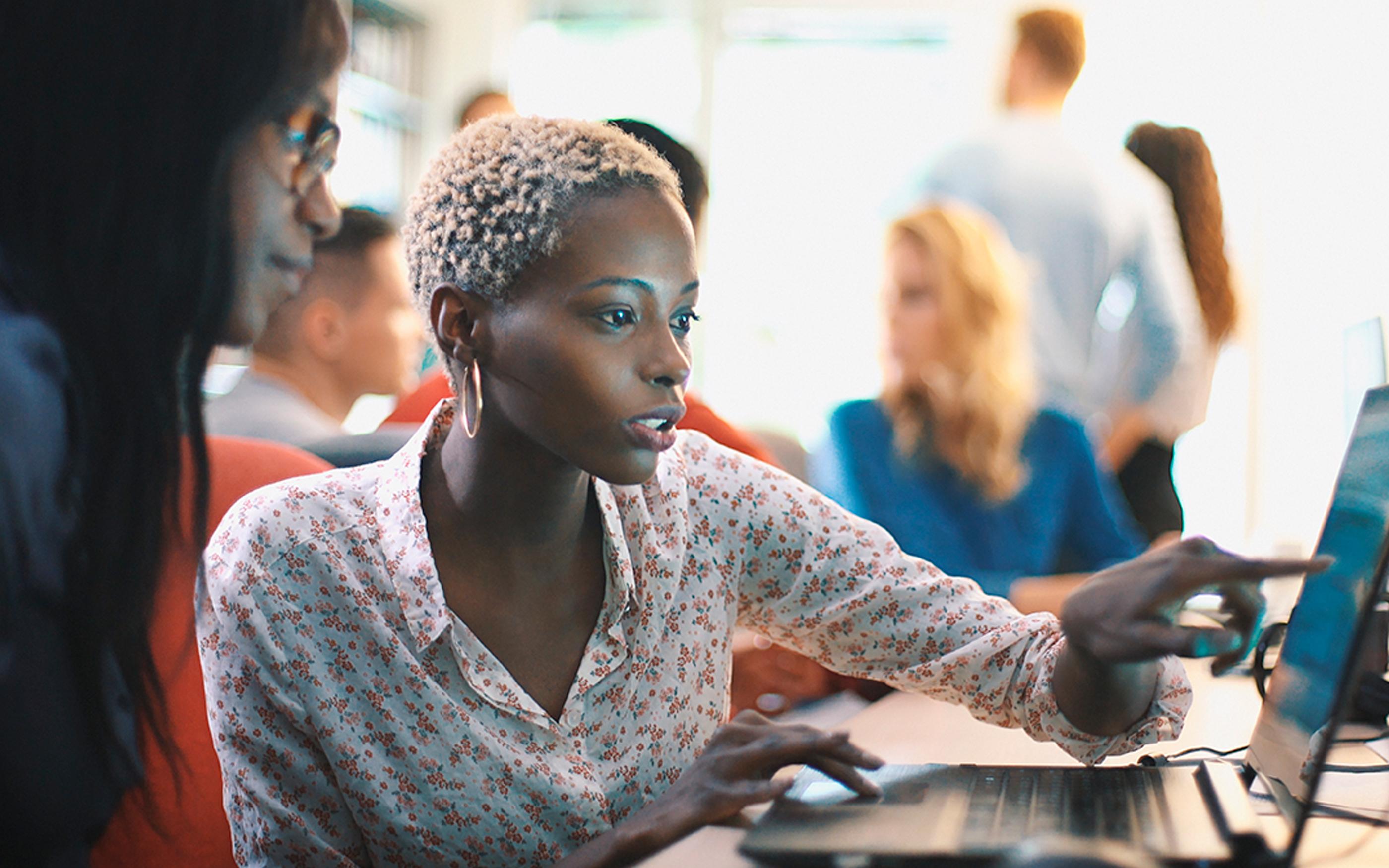 Two women colleagues working on the same laptop, with their co-workers in the background