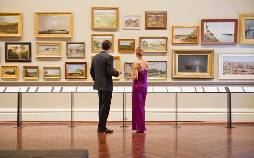 Two people in formal attire viewing an extensive art collection of framed landscape paintings on a gallery wall.