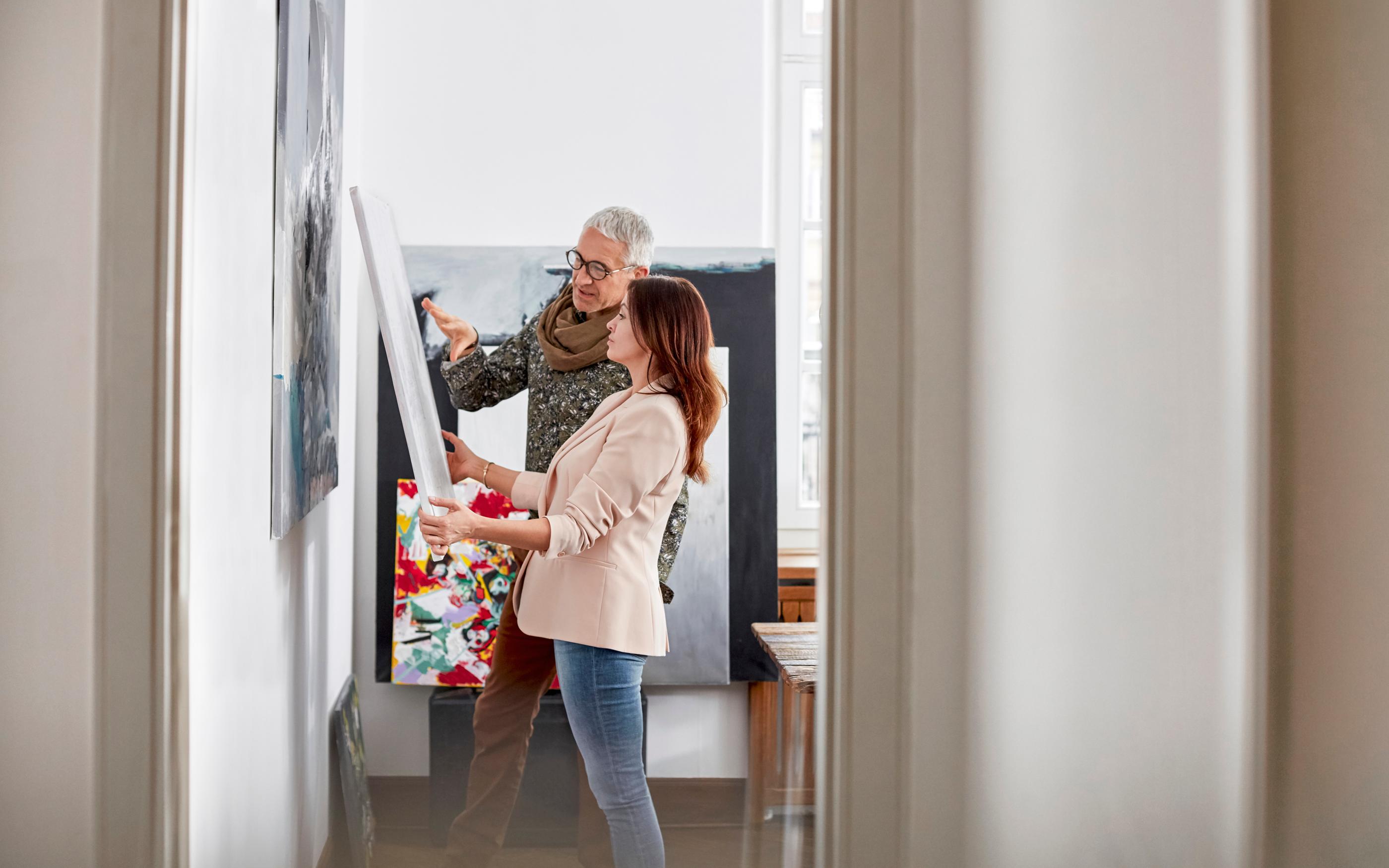 Two individuals carefully hanging a large piece of artwork on a wall in a clean, modern interior space.