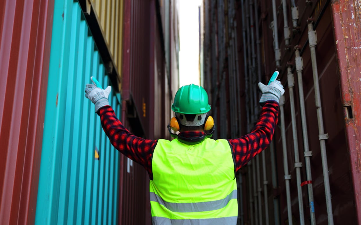A worker in a high-visibility vest and hard hat standing between large shipping containers with arms raised.